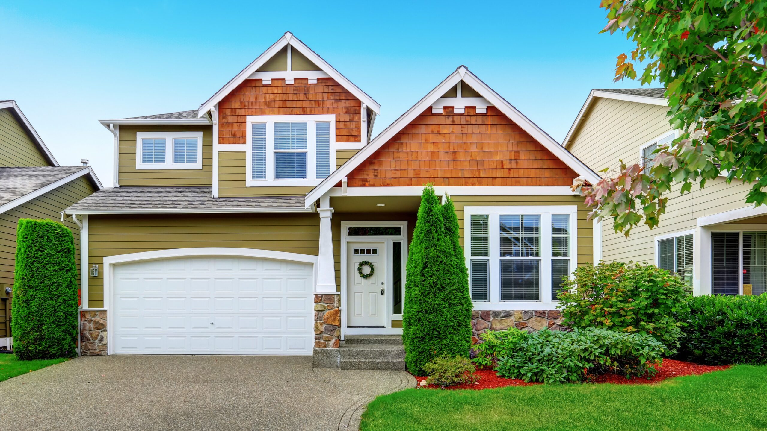 Small two-story American house and garage with olive exterior paint and freshly landscaped yard