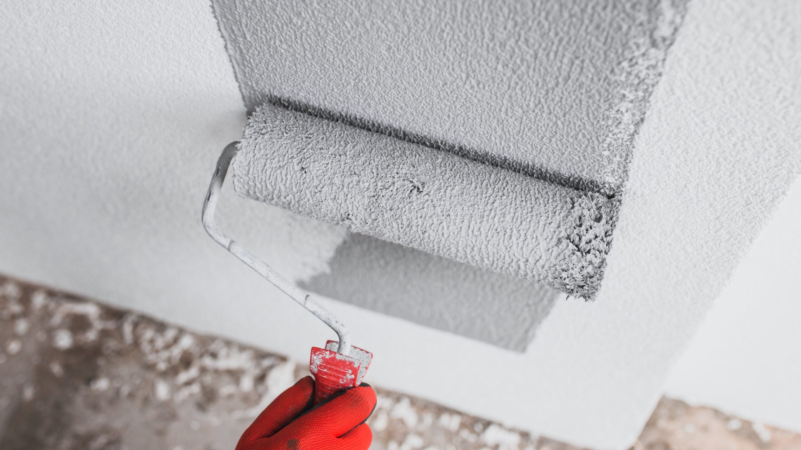 Red gloved hand using grey paint roller to paint the exterior of a building
