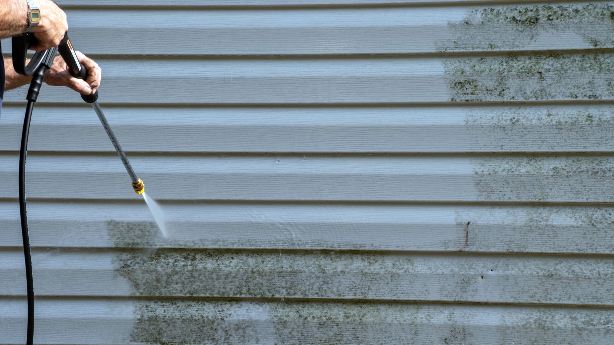 Two male hands using a pressure washer to spray off mold from white vinyl siding