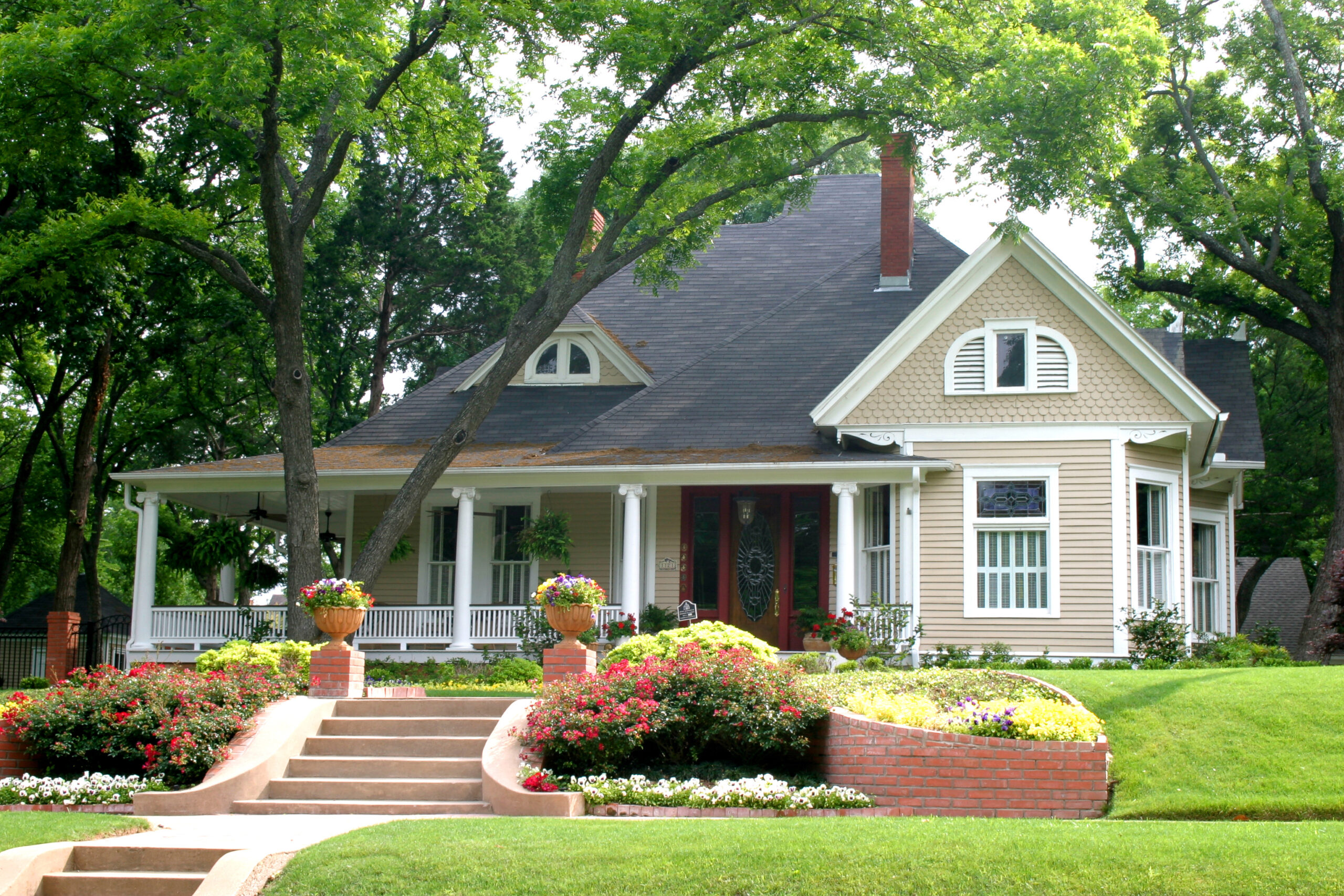 Tan house with brick red front door, and flower garden in front of it